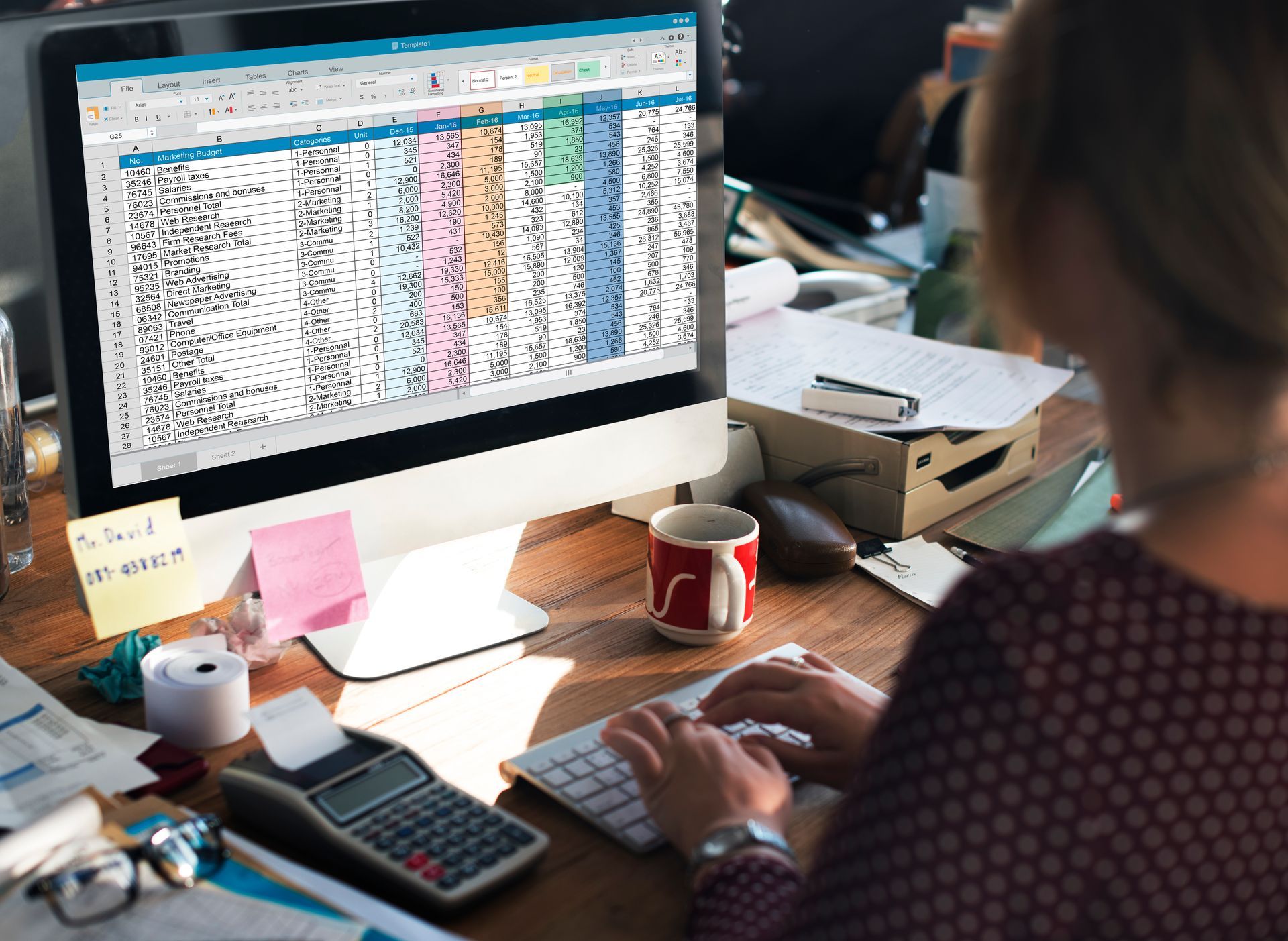 Woman working on a spreadsheet on a desktop computer at a desk, calculator, sticky notes, and coffee cup visible.