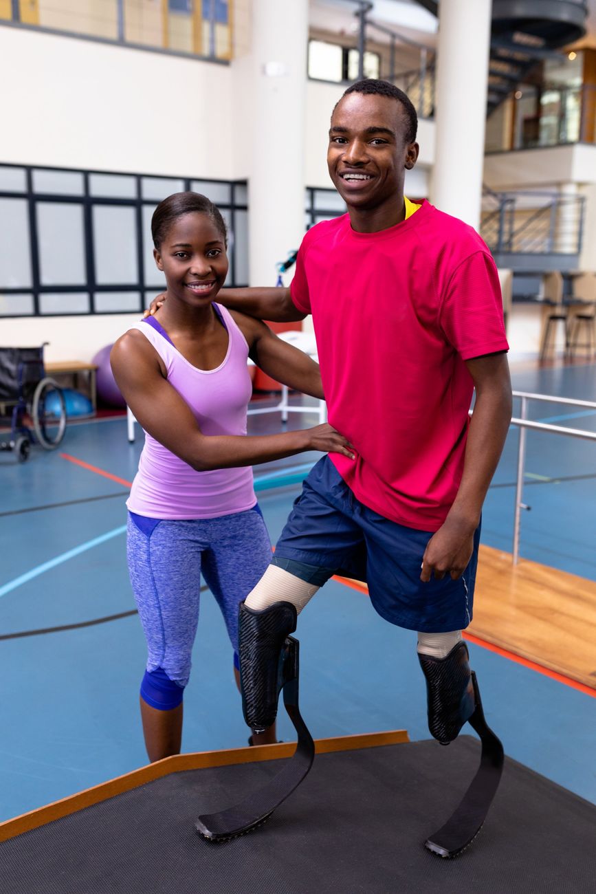 A man with prosthetic legs is being helped by a woman in a gym.