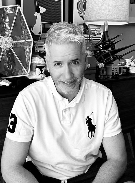 A man wearing a white polo shirt with a horse on it is smiling in a black and white photo.