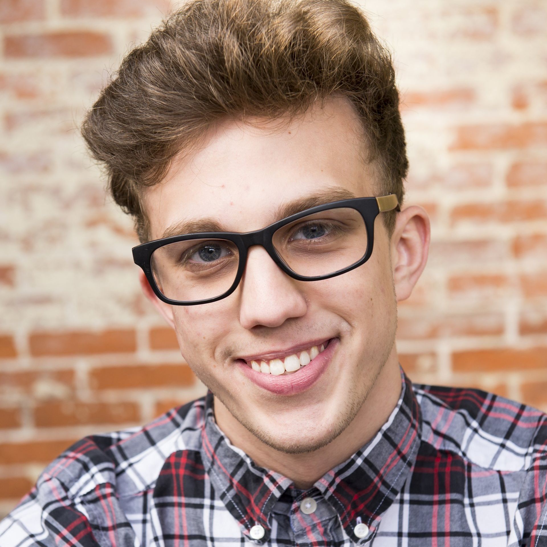 A young man wearing glasses and a plaid shirt is smiling in front of a brick wall.