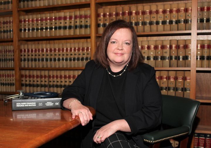 Woman in black blazer sits at a desk with law books in background.