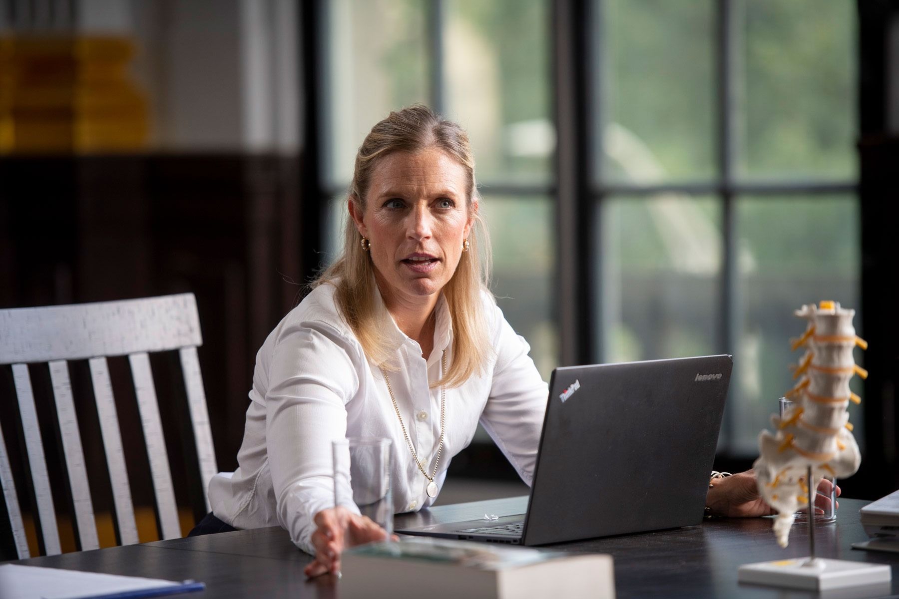 Woman at desk with laptop, gesturing; spine model, window, and books are present.