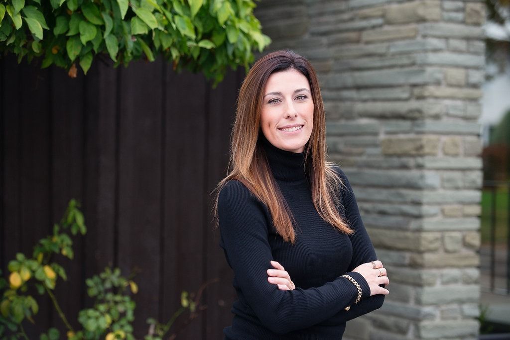 Woman with long brown hair in black turtleneck smiles, arms crossed, by a stone wall.