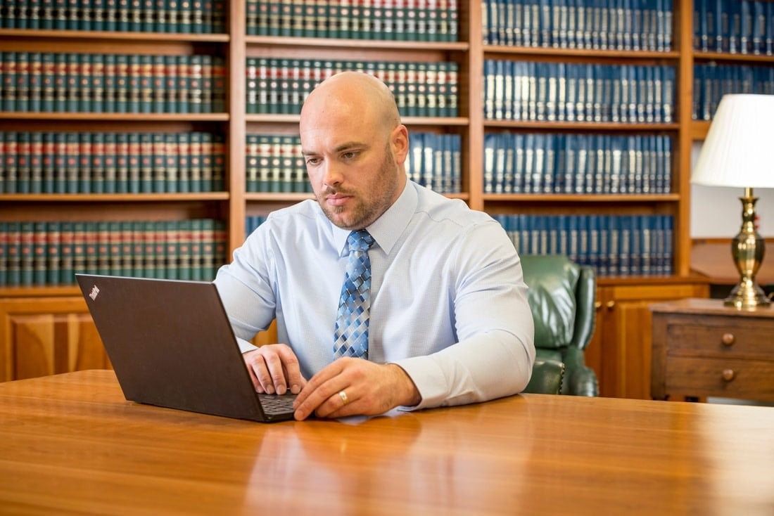 Bald man in a light blue shirt types on a laptop in a wood-paneled office with bookshelves.