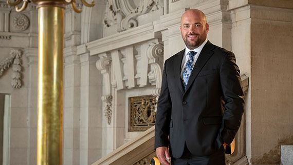 Man in a suit, smiling, standing in a grand building with ornate details, near a gold pillar.
