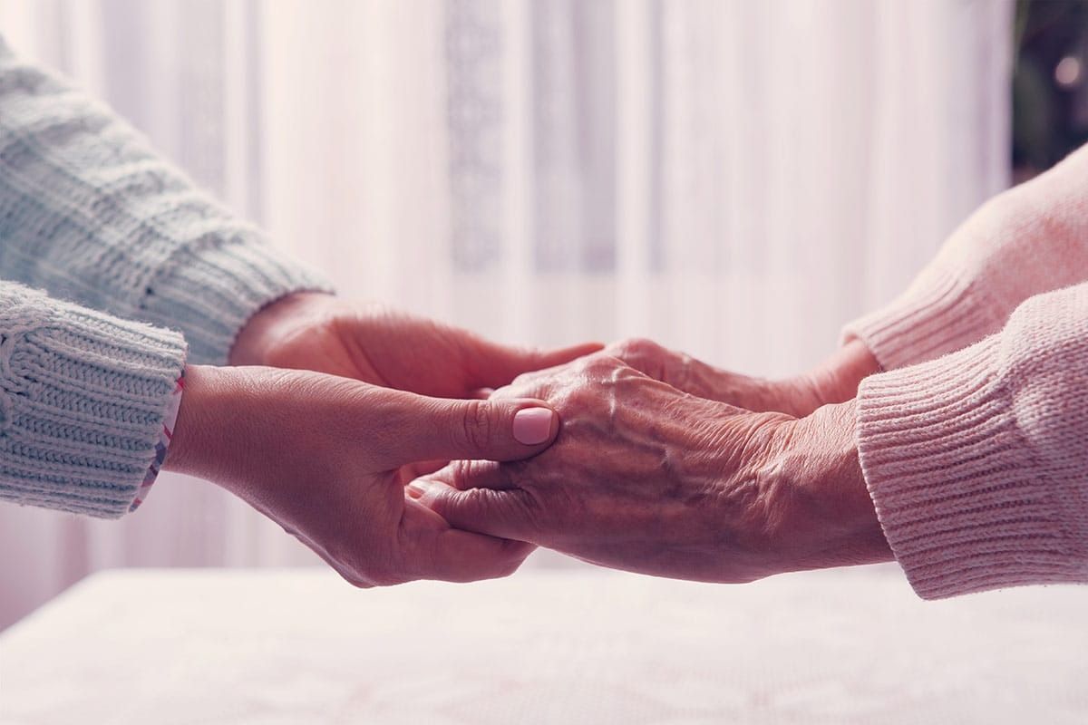 Two people holding hands; young person's hands over an older person's hands, offering support and care.