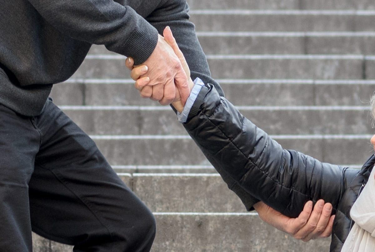 Man's hand grasping another person's arm, on a staircase. Grey sweater, black jacket, denim pants visible.