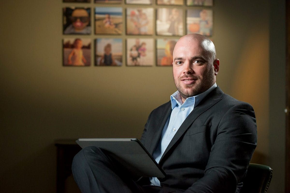 Man in suit, holding tablet, smiles at the camera. Photos on wall behind.