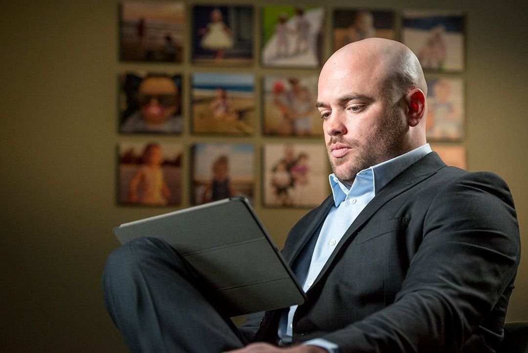 Bald man in suit, seated, looking at tablet, photos on wall behind him.