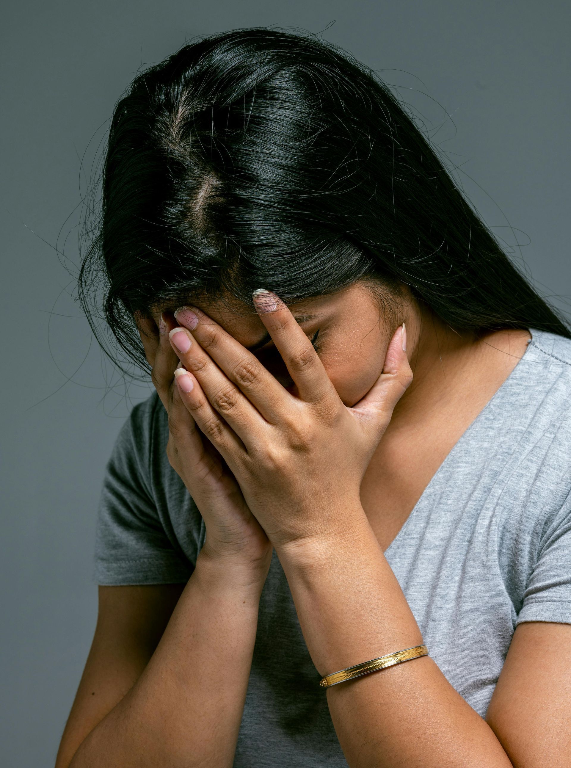 Woman with dark hair covering her face with her hands, looking upset, wearing a gray shirt and gold bracelet.