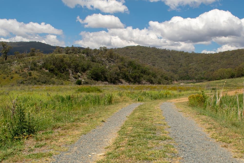 A Dirt Road Going Through A Grassy Field With Mountains — Shine Your Ride In Googong, NSW