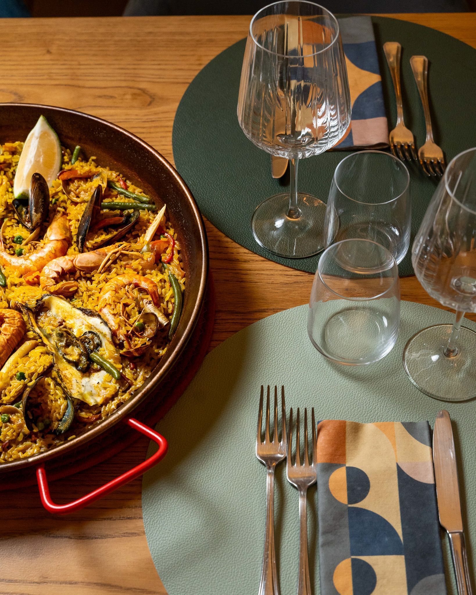 Paella su una tavola apparecchiata con bicchieri da vino, piatti e posate su tovagliette intrecciate.