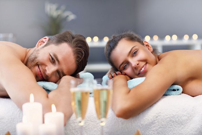 Due persone si rilassano in una spa, appoggiate ad asciugamani, con candele accese in primo piano.