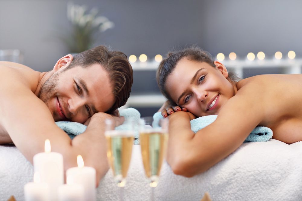 Due persone si rilassano in una vasca idromassaggio con candele accese, sorridendo e appoggiando la testa sugli asciugamani.