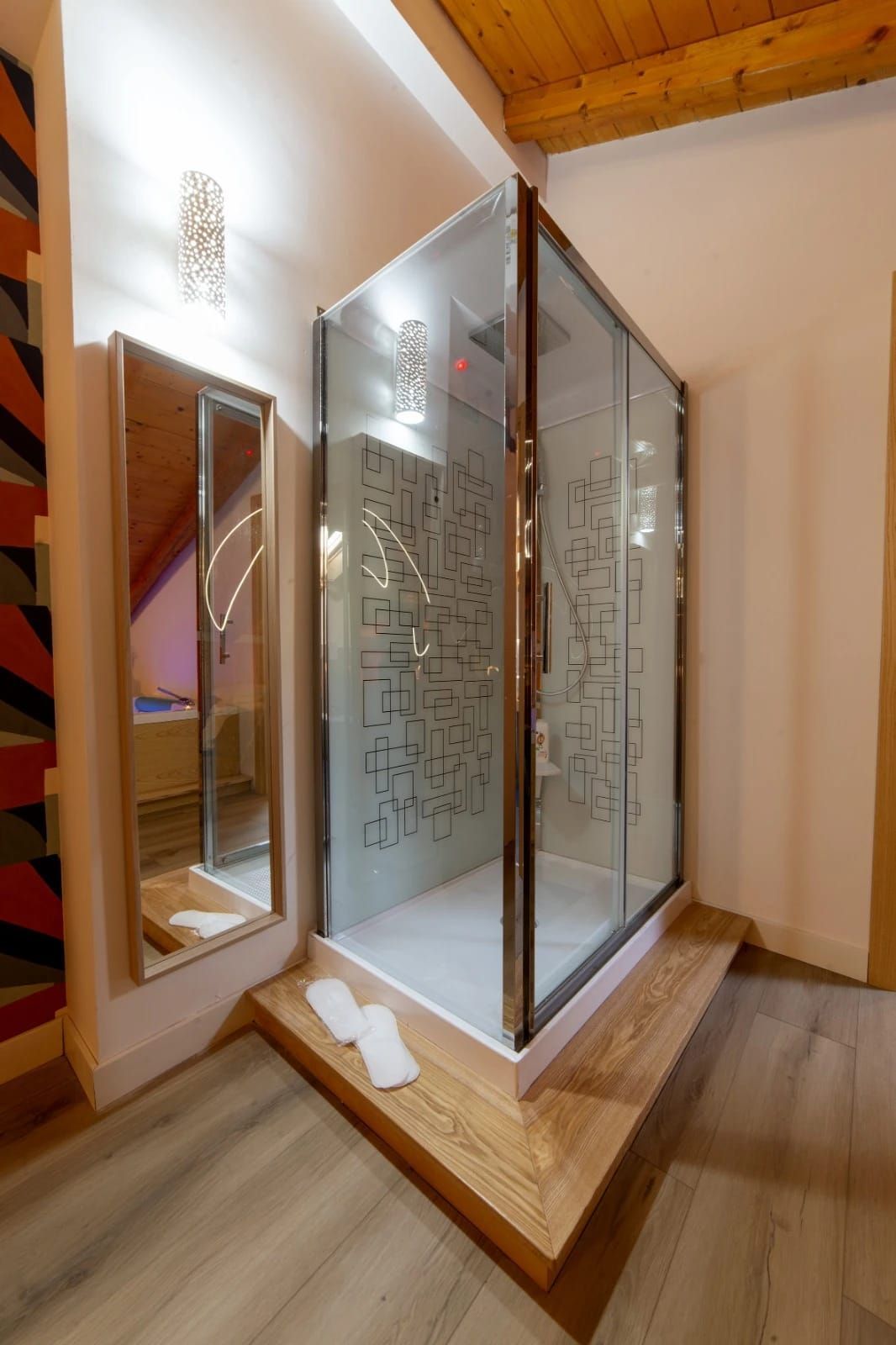 Box doccia moderno in vetro in un bagno con finiture in legno, parete a specchio e asciugamano sul pavimento.