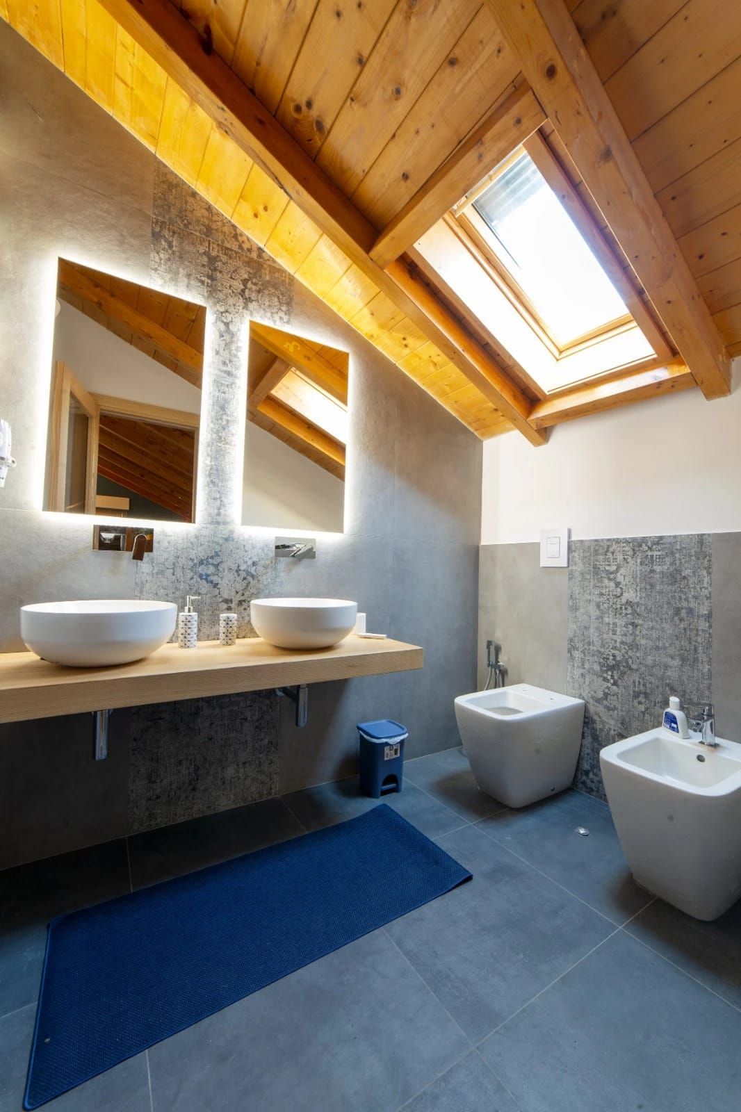Bagno moderno con lucernario con travi in legno, doppio lavabo, piastrelle grigie e sanitari bianchi.