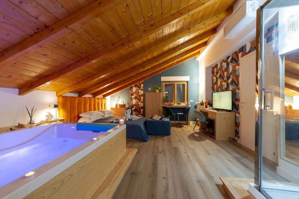 Accogliente camera da letto mansardata con vasca idromassaggio illuminata, soffitto in legno inclinato e zona relax vicino a grandi finestre.