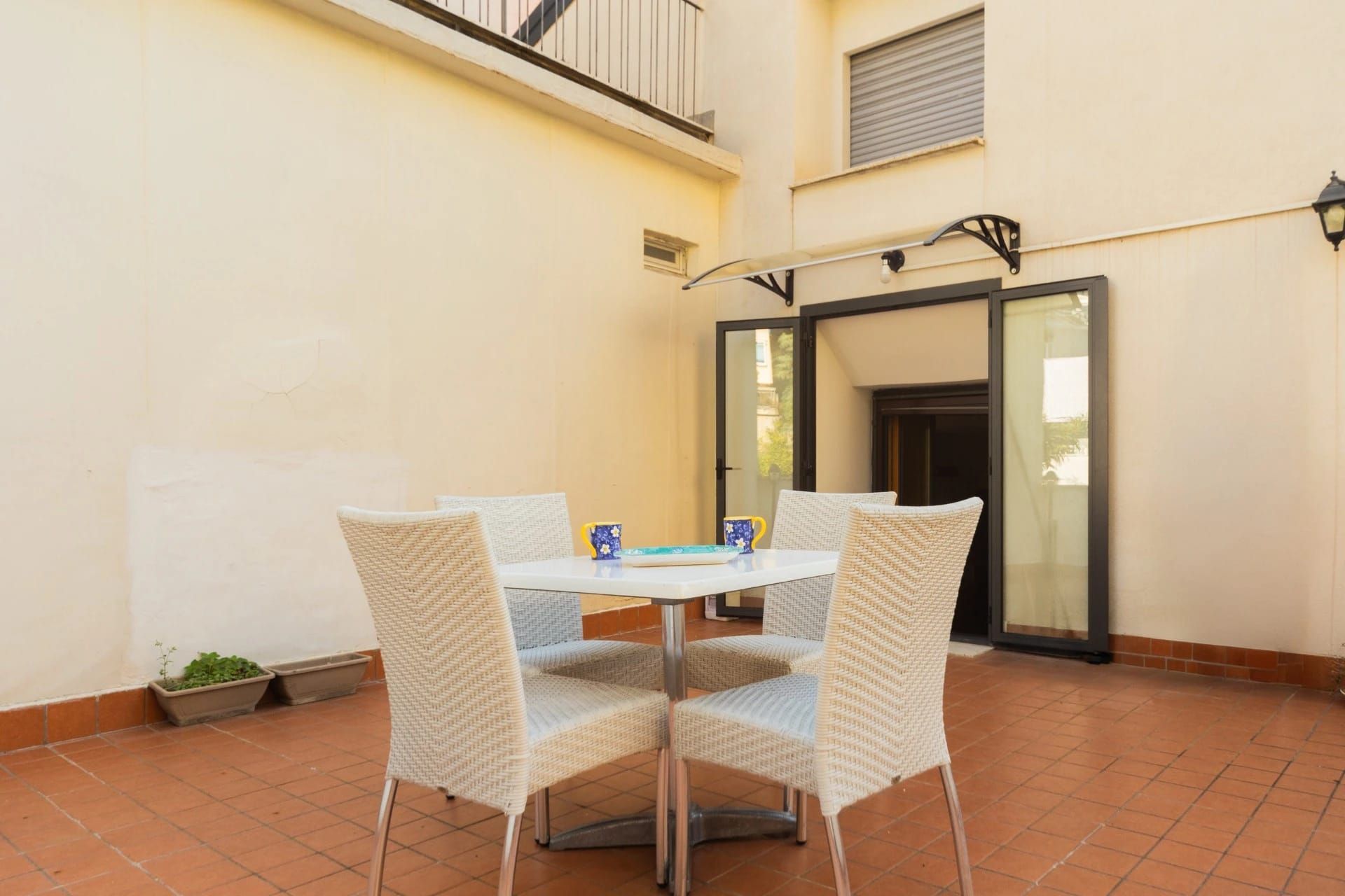 Patio con tavolo e sedie in vimini bianco su piastrelle di terracotta accanto a porte finestre aperte.