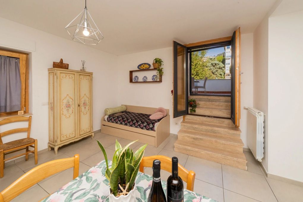Piccola camera da letto con armadio, letto, tavolo e porta che si apre su un balcone soleggiato.