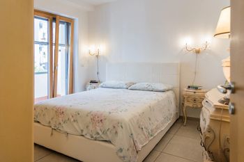 Camera da letto luminosa con letto bianco, biancheria da letto a motivi floreali, comodini e finestra con porte finestre che danno sul balcone.