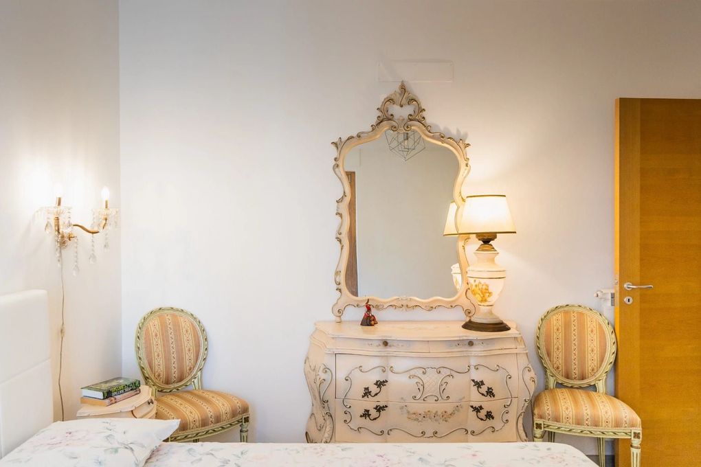 Elegante camera da letto con specchio decorato, comò, lampada e due sedie a righe su pareti bianche.