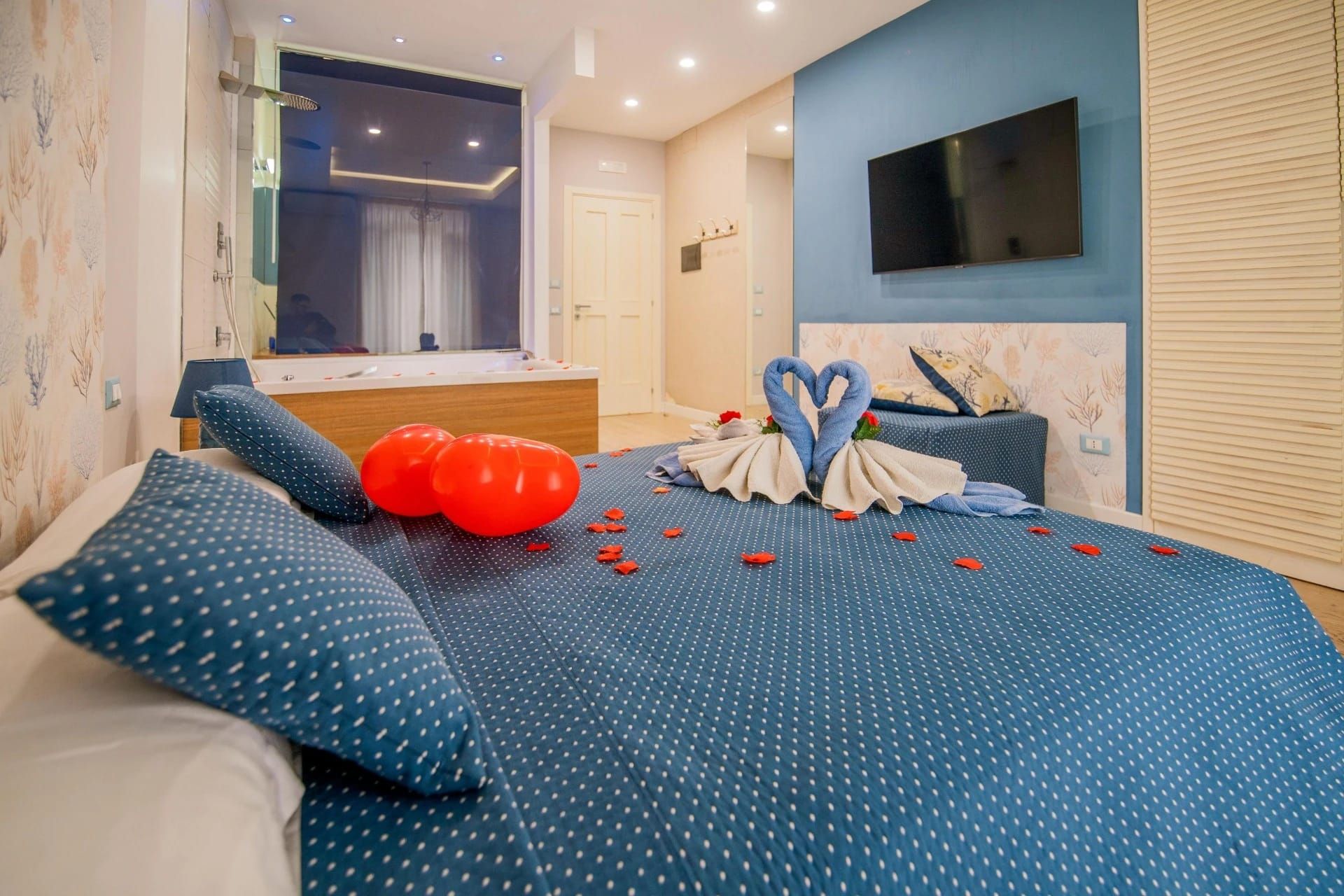 Letto a pois blu con petali di rosa, decorazioni a forma di cuore realizzate con asciugamani e palloncini rossi in una camera da letto moderna con TV.