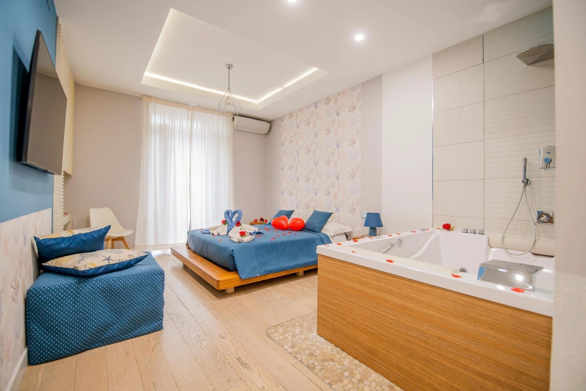 Luminosa camera da letto moderna con letti blu, TV a parete e vasca idromassaggio bianca vicino alla finestra.