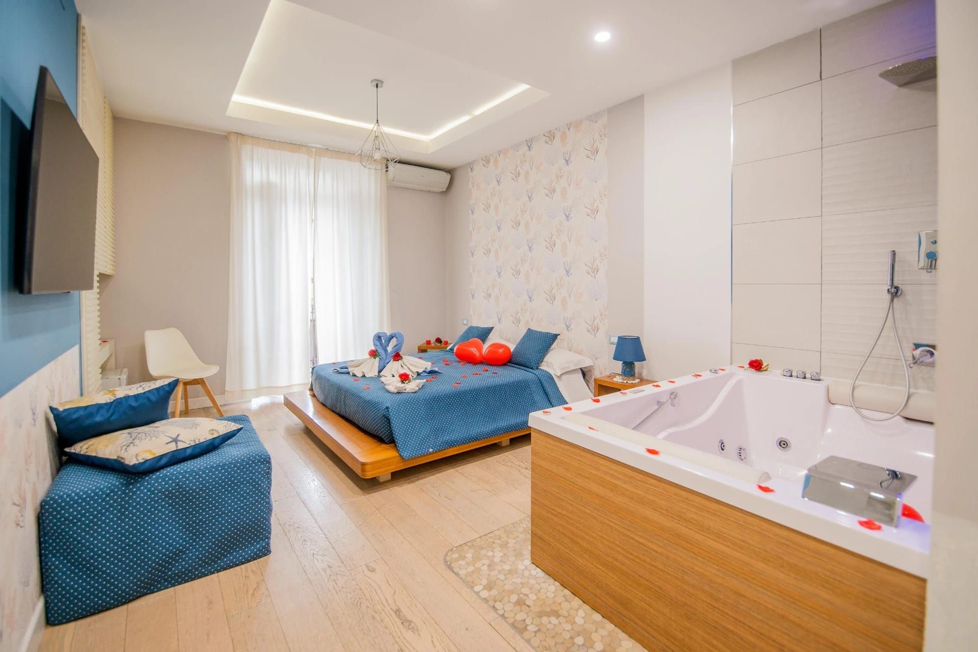 Camera da letto moderna con lenzuola blu, divano e vasca da bagno con pannelli in legno in una luminosa stanza bianca.