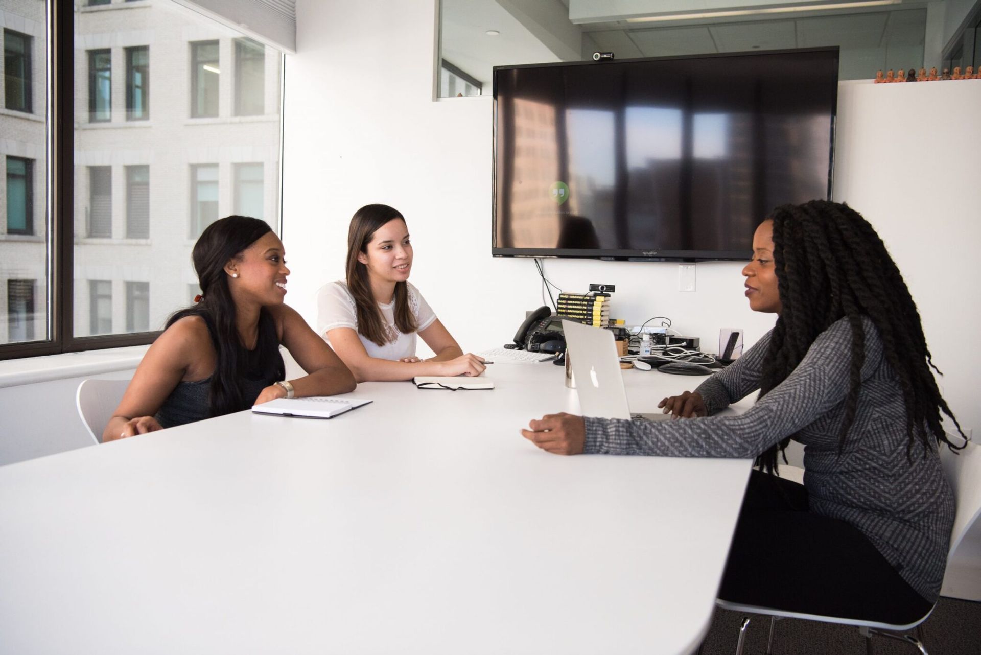 Three people in a meeting at a white table. One woman is speaking to the other two.
