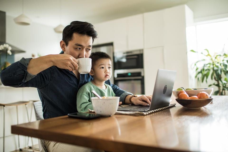 Man working on laptop, holding cup, child on lap with bowl of food; kitchen setting.