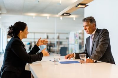 Woman gesturing, speaking with man at a table, documents and glasses of water present. Office setting with natural light.