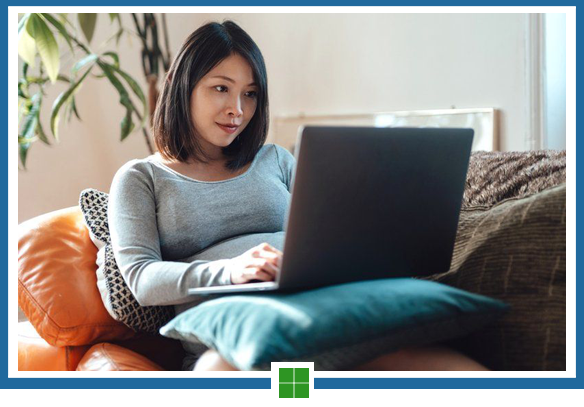 Woman using laptop, seated on a couch with pillows, indoors.