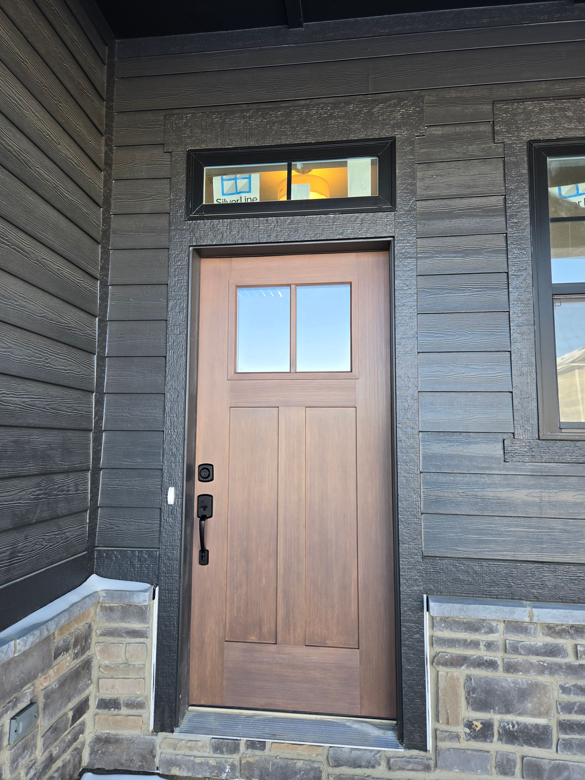 Brown door with glass panes, black siding, and a stone entryway.