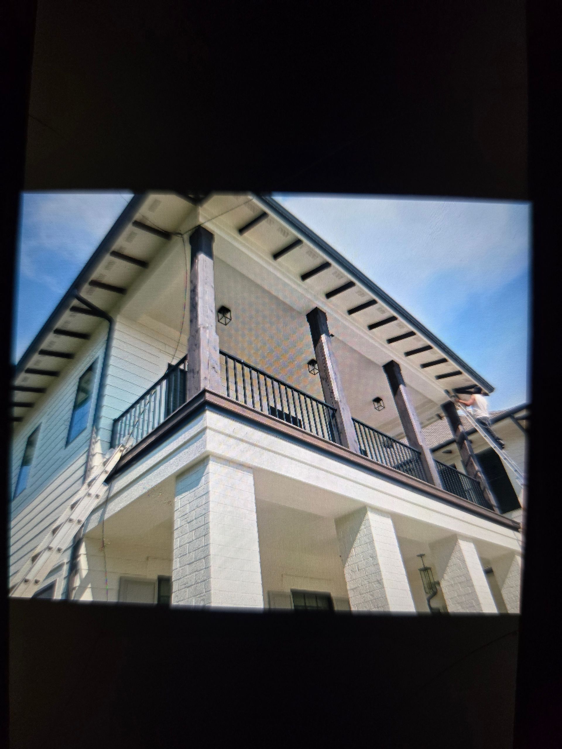 Two-story house with white columns, a dark roof, and a balcony with a black railing, set against a blue sky.
