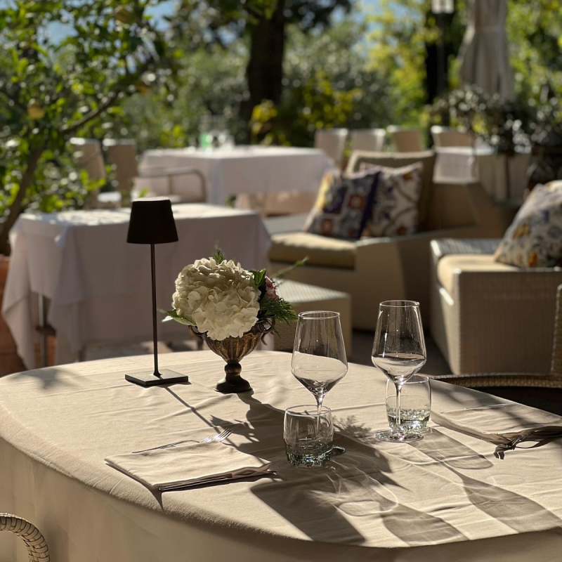 Tavolo da ristorante all'aperto apparecchiato con fiori, bicchieri e una lampada. Altri tavoli e sedute sullo sfondo. Giornata di sole.