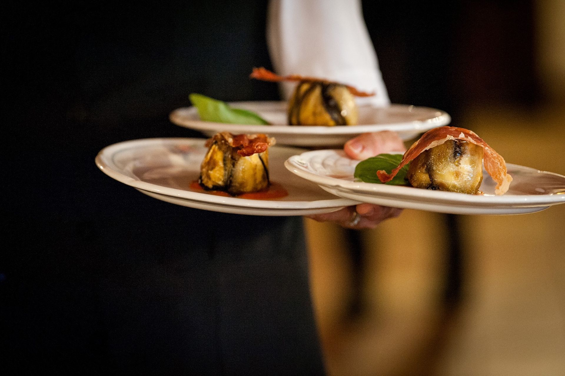 Cameriere che regge tre piatti di cibo; i piatti sembrano contenere melanzane e prosciutto.