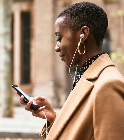 Une femme en manteau beige utilise son smartphone et écoute de la musique avec des écouteurs filaires à l'extérieur.