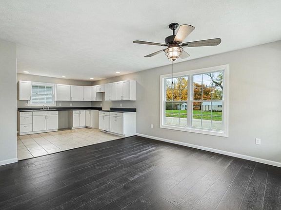 A living room with a ceiling fan and a kitchen in the background.
