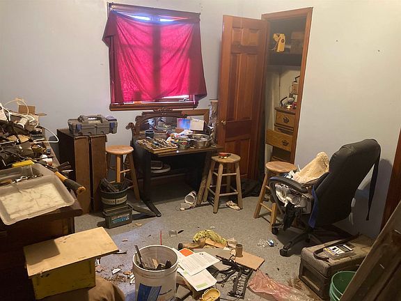 A messy room with a desk , chair , stools and a window.