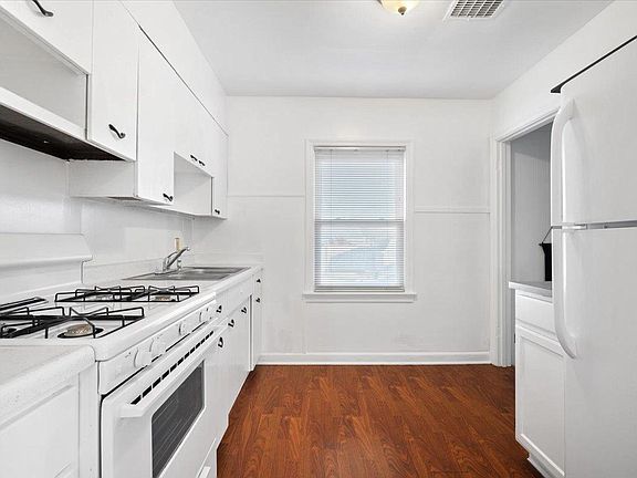 A kitchen with white cabinets , a stove , a refrigerator , and a window.