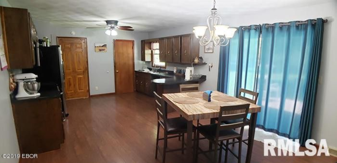 A dining room with a table and chairs and a kitchen in the background.