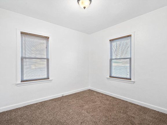 An empty bedroom with two windows and a brown carpet.