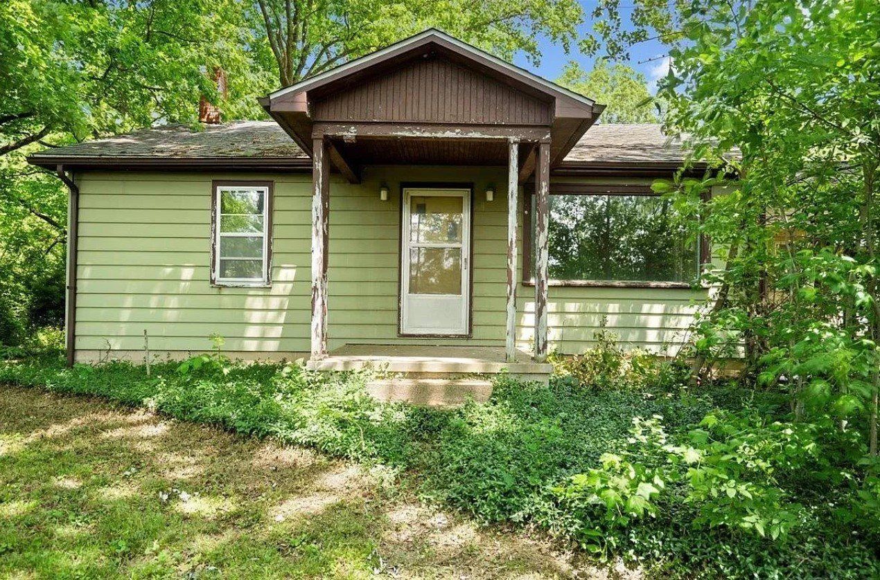 A small green house with a porch is surrounded by trees.