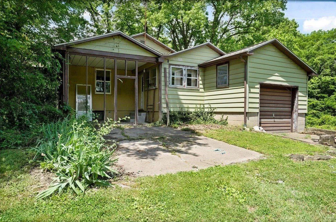 A small house with a porch and a garage is surrounded by trees.