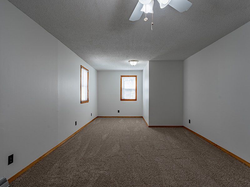 An empty living room with a ceiling fan and two windows.