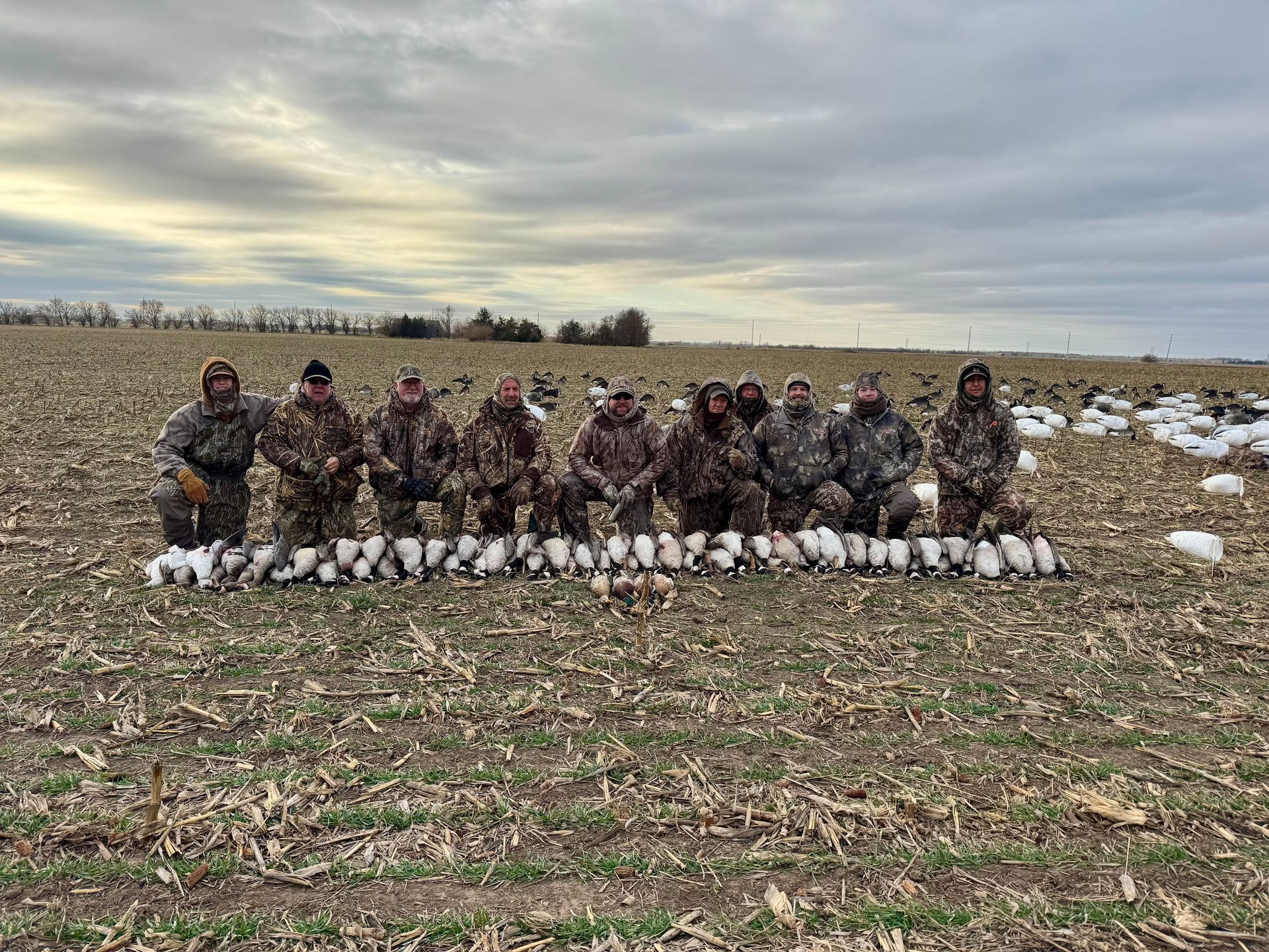 Group of hunters with camo gear, pose with harvested geese in a field, under cloudy sky.