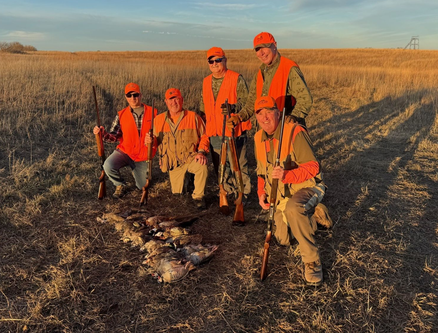 Five people in orange vests pose with hunting rifles and dead birds in a field.