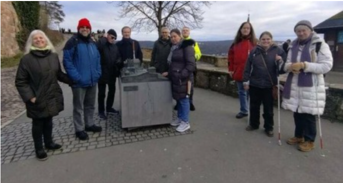 Eine Gruppe von Menschen, die sich im Freien versammelt haben, einige tragen Winterkleidung. In der Mitte steht ein Modell oder eine Skulptur, um die sich die Gruppe versammelt hat. Einige der Personen scheinen weiße Blindenstöcke zu benutzen, was darauf hindeutet, dass sie sehbehindert sind. Im Hintergrund ist eine Mauer sowie eine winterliche Landschaft zu sehen.