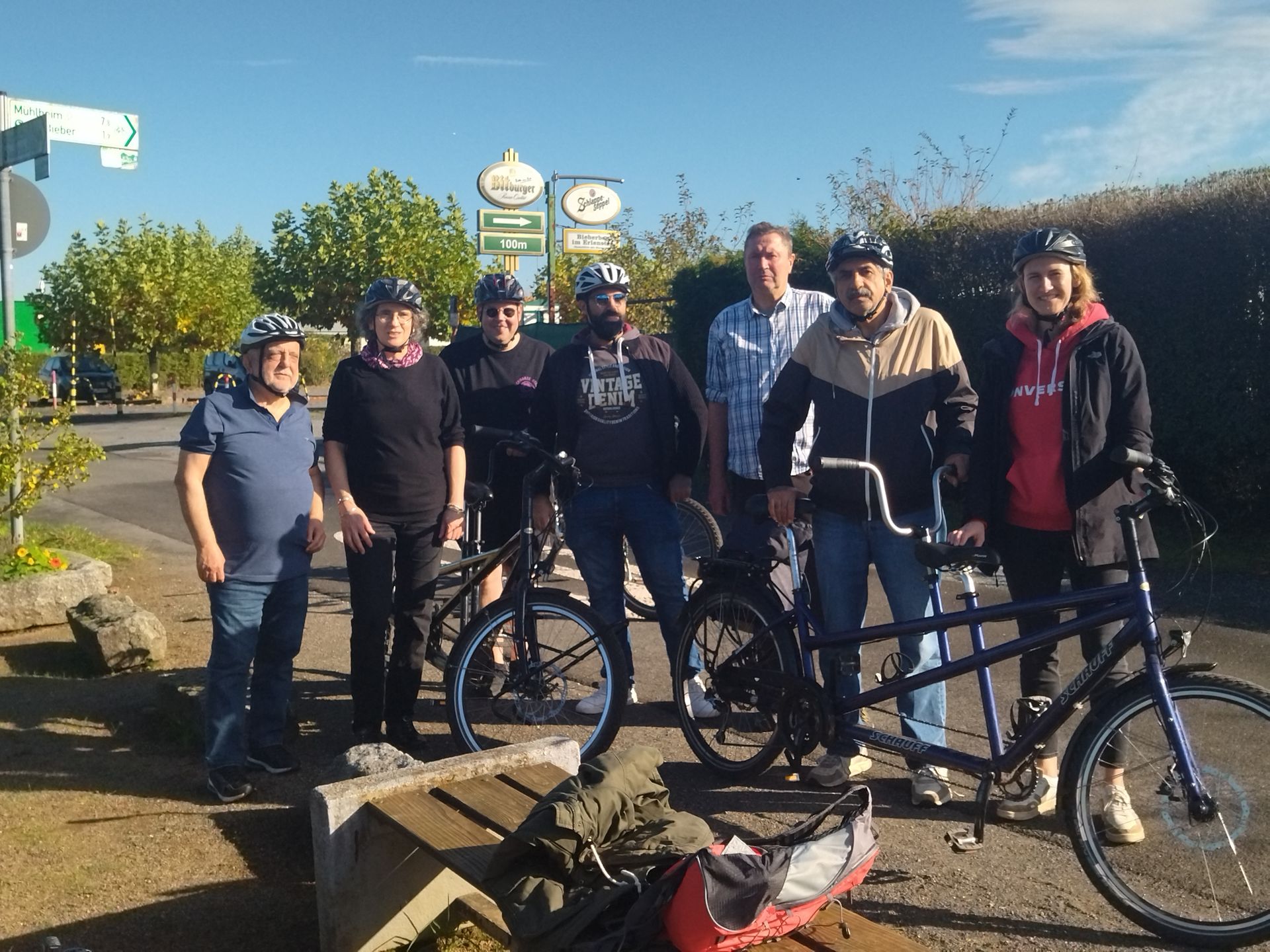 Eine Gruppe von sieben Personen steht mit Fahrrädern, darunter ein Tandem, auf einem Platz bei sonnigem Wetter. Alle tragen Fahrradhelme außer eine Person, die in Freizeitkleidung gekleidet ist. Im Hintergrund sind grüne Büsche, ein Wegweiser und Schilder eines Restaurants oder Cafés zu sehen. Die Gruppe wirkt freundlich und entspannt. Im Vordergrund liegt eine Tasche auf einer Bank.