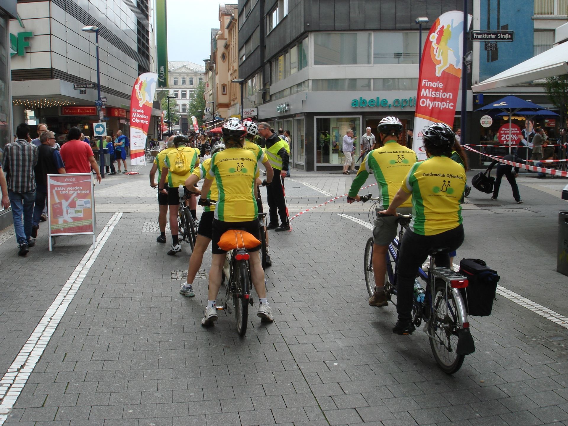 Eine Gruppe von Menschen fährt mit dem Fahrrad eine Straße entlang vor einem Geschäft, auf dem „Siemens“ steht